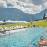 Ein Mann und eine Frau sitzen am Rande eines Außenpools mit Liegestühlen und Sonnenschirmen, umgeben von grünen Feldern und Bergen unter einem teilweise bewölkten Himmel.