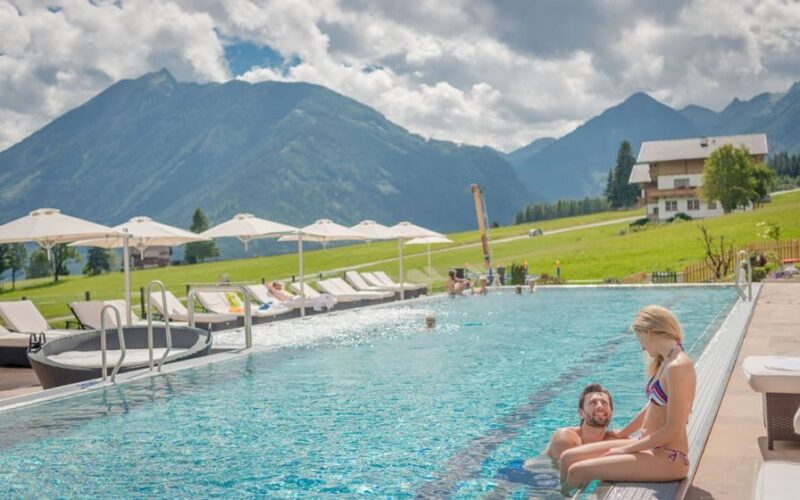 Ein Mann und eine Frau sitzen am Rande eines Außenpools mit Liegestühlen und Sonnenschirmen, umgeben von grünen Feldern und Bergen unter einem teilweise bewölkten Himmel.