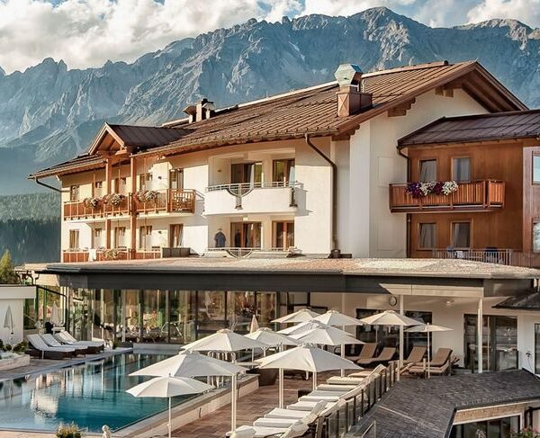 Ein modernes Alpenhotel mit Balkonen und einem Poolbereich, umgeben von Liegestühlen und Sonnenschirmen, vor dem Hintergrund bewaldeter Hügel und schroffer Berge.