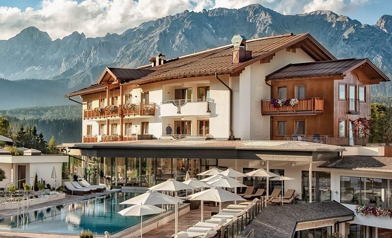 Ein modernes Alpenhotel mit Balkonen und einem Poolbereich, umgeben von Liegestühlen und Sonnenschirmen, vor dem Hintergrund bewaldeter Hügel und schroffer Berge.
