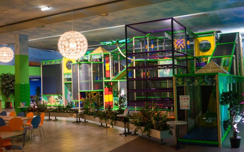 Indoor-Spielplatz mit farbenfrohen Klettergerüsten und Rutschen, umgeben von Pflanzen und einem Sitzbereich mit Tischen und Stühlen, unter hängenden dekorativen Lampen.