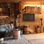 Ein rustikaler Holzraum mit Fässern, Wannen, hängenden Töpfen und Küchengeräten, mit Steinmetzarbeiten um einen großen Kessel und Regalen mit runden Holztabletts.