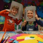 Zwei Kinder sitzen an einem mit buntem Papier und Bastelmaterial bedeckten Tisch, halten sich Papprahmen vors Gesicht und lächeln in die Kamera. An der Wand hinter ihnen ist ein Graffiti-Wandbild zu sehen.