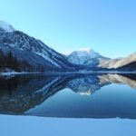 In einem ruhigen See spiegeln sich schneebedeckte Berge und Bäume unter einem klaren blauen Himmel wider, im Vordergrund ist das Ufer mit Schnee bedeckt.