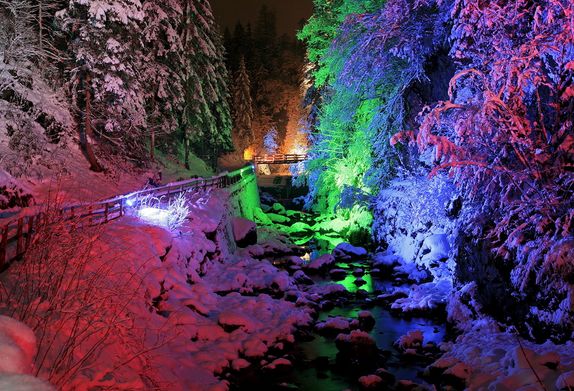 Ein verschneiter Waldbach wird nachts von bunten Lichtern erleuchtet, die Schnee, Bäume und Wasser in rote, grüne, blaue und violette Farbtöne tauchen.