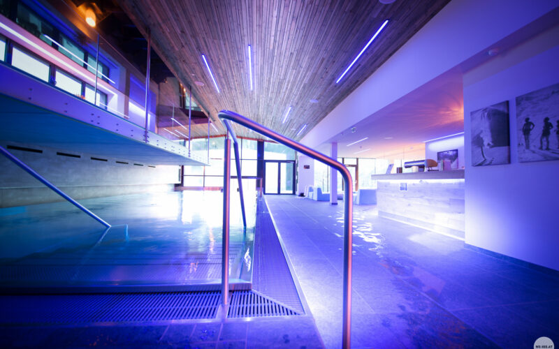 Moderner Hallenbadbereich mit blauer Beleuchtung, Holzdecke, Metallgeländern und einem Empfangstresen im Hintergrund.
