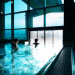 Zwei Personen entspannen sich in einem Hallenbad neben großen Fenstern, während draußen eine verschneite Berglandschaft zu sehen ist.