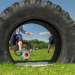 Eine Person kickt einen Fußball durch einen großen Traktorreifen, der im Freien auf dem Rasen liegt, während im Hintergrund andere Personen zu sehen sind.