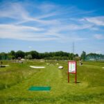 Ein grasbewachsener Discgolfplatz unter blauem Himmel mit vereinzelten Wolken, mit einem Abschlagplatz, Schildern, Sandfängen und Menschen in der Ferne.