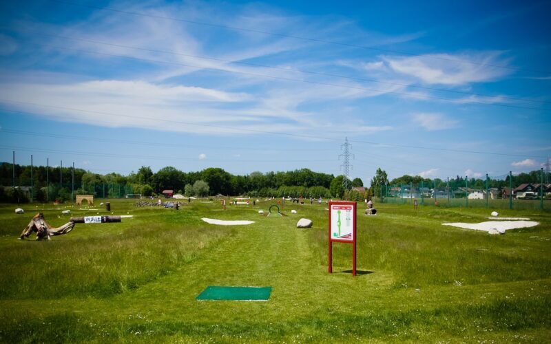 Ein grasbewachsener Discgolfplatz unter blauem Himmel mit vereinzelten Wolken, mit einem Abschlagplatz, Schildern, Sandfängen und Menschen in der Ferne.