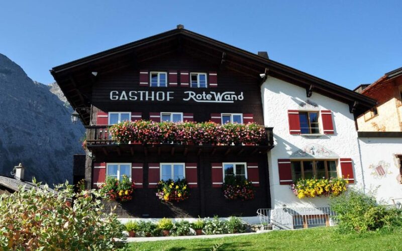 Ein traditionelles Alpengasthaus mit Blumenkästen unter den Fenstern und dem Schriftzug "Gasthof Rote Wand" an der Fassade, vor einem strahlend blauen Himmel.