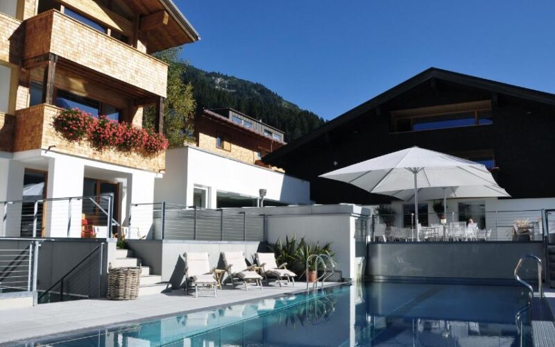 Modernes Gebirgsresort mit Holzbalkonen, Liegestühlen und einem großen weißen Sonnenschirm neben einem klaren Außenpool unter blauem Himmel.