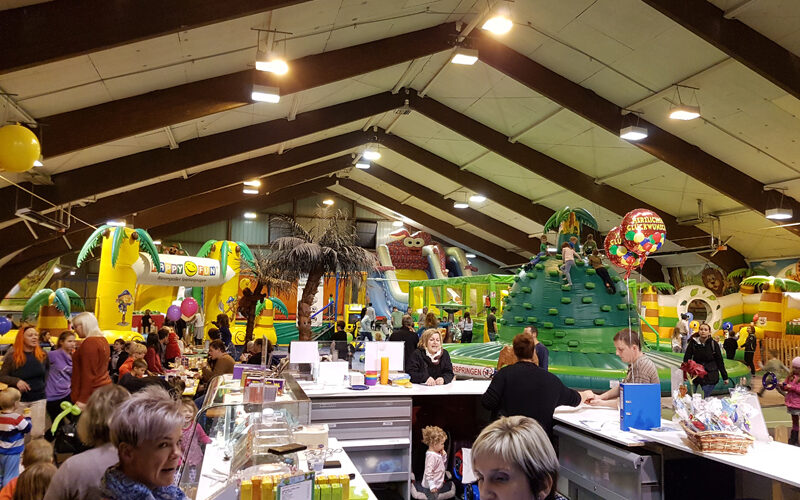 Indoor-Spielplatz mit Hüpfburgen und Rutschen, belebt von Kindern und Erwachsenen. Im Vordergrund befindet sich ein Thekenbereich, in dem Speisen und Waren angeboten werden, und Mitarbeiter, die sich um die Kunden kümmern.