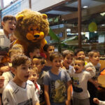 Eine Gruppe Kinder posiert und lächelt mit einer Person in einem Löwenkostüm in einem hell erleuchteten Innenraum.