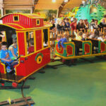 Kinder fahren in einem Vergnügungszentrum mit einer kleinen Indoor-Eisenbahn, während Erwachsene und andere Kinder hinter einem Zaun zuschauen.