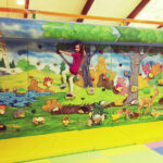 Ein Kind in einem roten Outfit klettert eine bunte Indoor-Kletterwand mit einem Wald- und Burgwandbild über gelben Polstermatten hoch.
