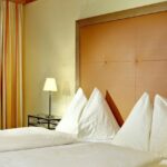 Ein ordentlich gemachtes Hotelbett mit vier dreieckigen weißen Kissen, einem orangefarbenen gepolsterten Kopfteil, einer Nachttischlampe und gestreiften Vorhängen am Fenster.