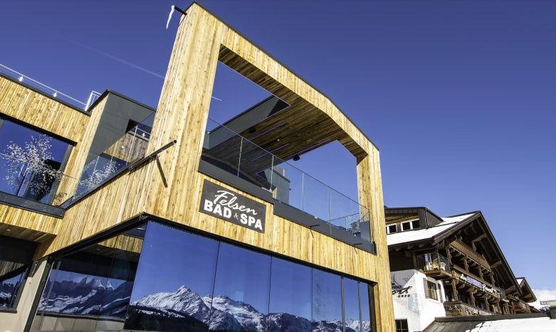 Modernes hölzernes Kurhaus mit großen Glasfenstern, in denen sich schneebedeckte Berge spiegeln, unter einem klaren blauen Himmel. Auf einem Schild steht "Felsen BAD + SPA".