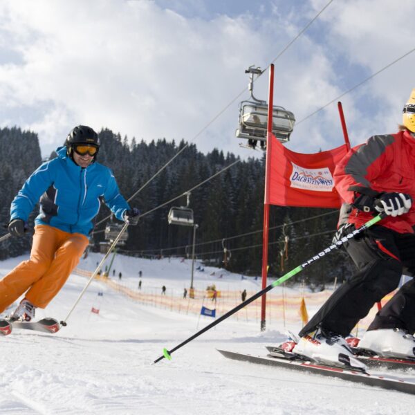 Zwei Personen fahren auf einer verschneiten Piste mit Skiliften und Kiefern im Hintergrund bergab; die eine trägt rot, die andere blau und orange.