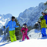Vier Personen in farbenfroher Skiausrüstung stehen an einem verschneiten Berghang, umgeben von Bäumen und Bergen, unter einem klaren blauen Himmel.