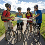 Vier Radfahrer stehen auf einem gepflasterten Weg in einer ländlichen Gegend und schauen gemeinsam auf eine Karte. Sie tragen Helme und Sportkleidung, im Hintergrund sind Felder, Hügel und Schilder zu sehen.