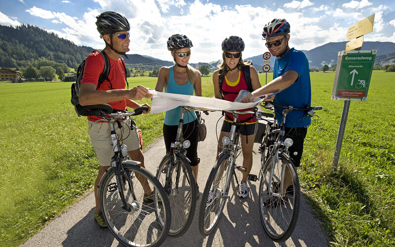 Vier Radfahrer stehen auf einem gepflasterten Weg in einer ländlichen Gegend und schauen gemeinsam auf eine Karte. Sie tragen Helme und Sportkleidung, im Hintergrund sind Felder, Hügel und Schilder zu sehen.