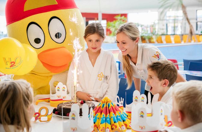 Kinder und ein Erwachsener versammeln sich um eine Geburtstagstorte mit Wunderkerzen; eine Person in einem großen gelben Vogelkostüm steht hinter ihnen. Auf dem Tisch sind Partydekorationen und Hüte zu sehen.