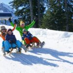 Zwei Erwachsene und zwei Kinder rodeln einen verschneiten Hügel hinunter, mit Bäumen und Bergen im Hintergrund, alle tragen Winterkleidung und lächeln.