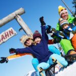 Eine winterlich gekleidete Familie sitzt auf Schlitten unter einem Holzschild mit der Aufschrift "RODELBAHN", lächelt und hebt an einem sonnigen Tag die Arme in den Schnee.