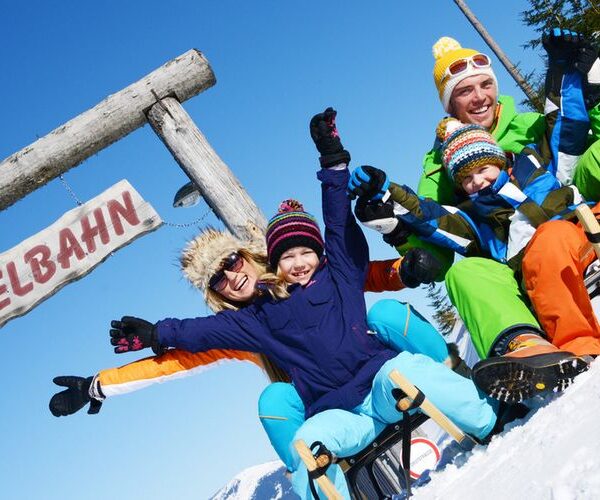 Eine winterlich gekleidete Familie sitzt auf Schlitten unter einem Holzschild mit der Aufschrift "RODELBAHN", lächelt und hebt an einem sonnigen Tag die Arme in den Schnee.