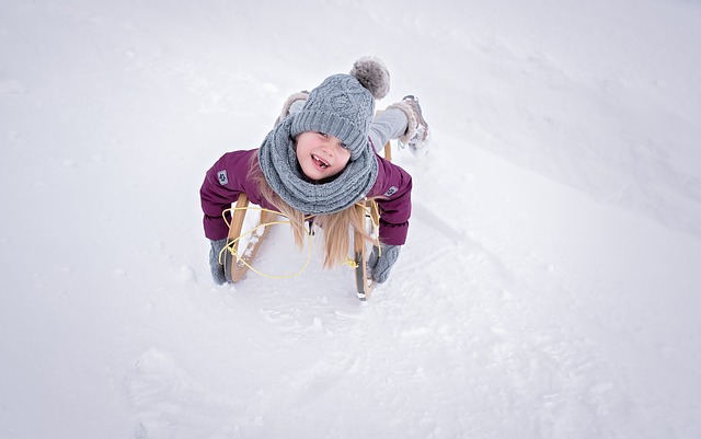 Ein winterlich gekleidetes Kind liegt mit dem Gesicht nach unten auf einem Holzschlitten im Schnee, schaut nach oben und lächelt.