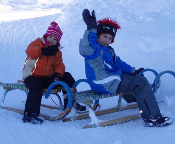 Zwei Kinder mit Winterjacken und Mützen sitzen auf Schlitten im Schnee und winken der Kamera zu.