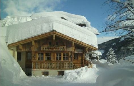 Ein Holzchalet mit einer dicken Schneeschicht auf dem Dach und dem umliegenden Boden, eingebettet in eine verschneite Berglandschaft unter blauem Himmel.