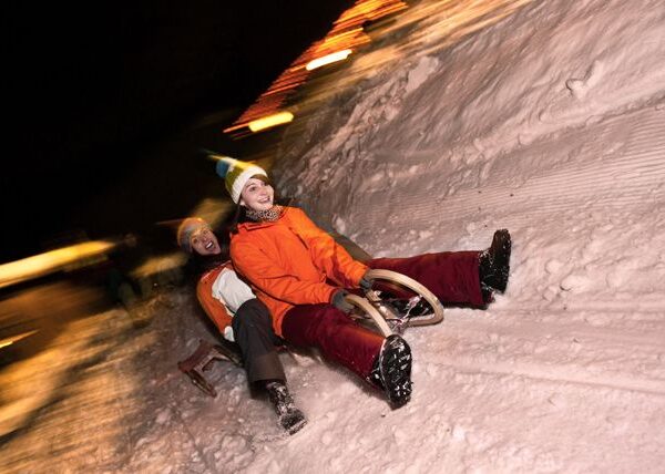 Zwei Personen in Winterkleidung fahren nachts mit einem Schlitten einen verschneiten Hügel hinunter, wobei verschwommene Lichter im Hintergrund die Bewegung anzeigen.