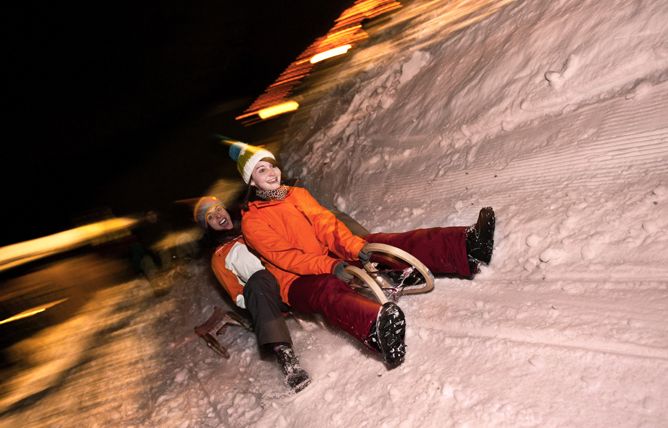 Zwei Personen in Winterkleidung fahren nachts mit einem Schlitten einen verschneiten Hügel hinunter, wobei verschwommene Lichter im Hintergrund die Bewegung anzeigen.