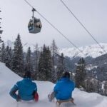 Zwei Personen in Winterjacken rodeln einen verschneiten Hügel hinunter, während über ihnen eine Seilbahn vorbeifährt und im Hintergrund schneebedeckte Berge zu sehen sind.