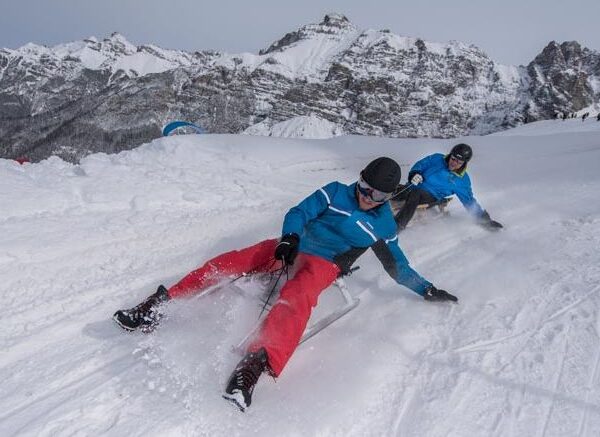 Zwei Personen in Winterkleidung fahren mit Schlitten einen verschneiten Hang hinunter, mit Bergen im Hintergrund.