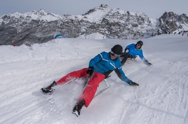 Zwei Personen in Winterkleidung fahren mit Schlitten einen verschneiten Hang hinunter, mit Bergen im Hintergrund.