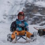 Ein Junge mit Helm und Winterausrüstung fährt auf einem Schlitten einen verschneiten Hang hinunter, während ein anderes Kind im Hintergrund vom Schlitten fällt.