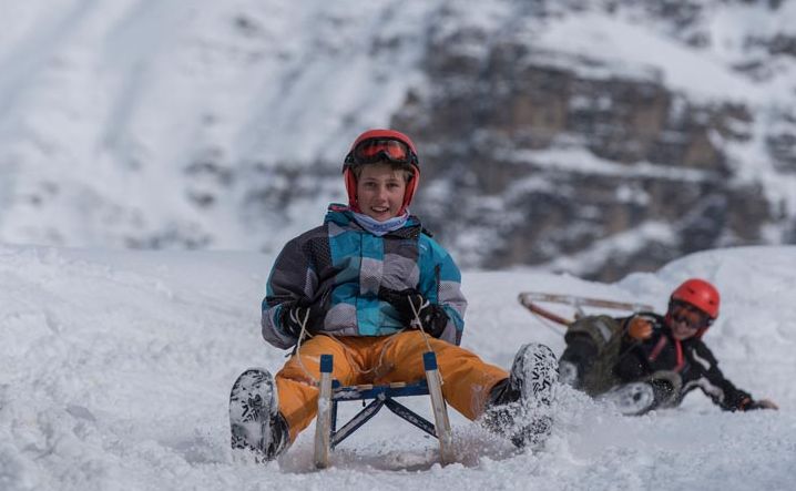 Ein Junge mit Helm und Winterausrüstung fährt auf einem Schlitten einen verschneiten Hang hinunter, während ein anderes Kind im Hintergrund vom Schlitten fällt.