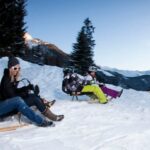 Drei Personen fahren auf Holzschlitten einen verschneiten Hang hinunter, im Hintergrund sind schneebedeckte Bäume und Berge zu sehen.