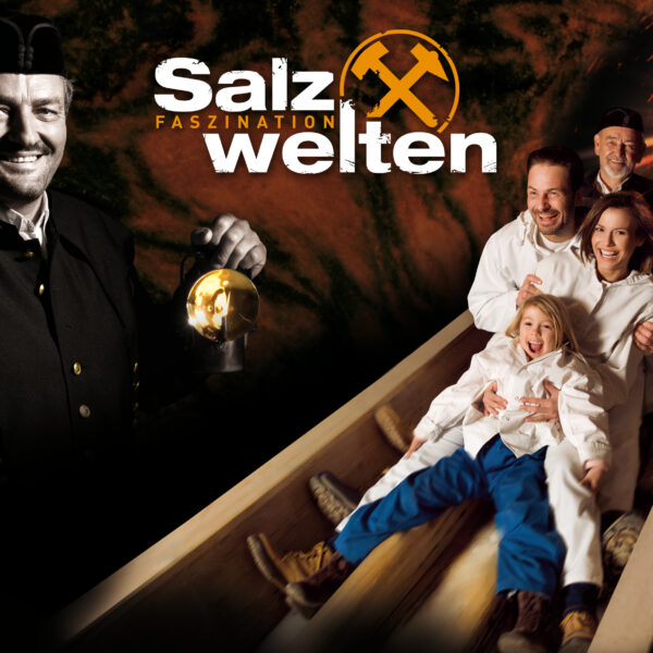 Eine Gruppe von Menschen in weißen Anzügen gleitet in einem Salzbergwerk eine Holzrutsche hinunter. Ein Bergmann hält eine Laterne und den Text „Salzwelten Faszination“ in der Hand.