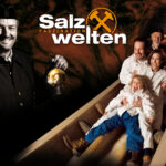 Ein lächelnder Mann in Bergmannsuniform hält eine Lampe, während eine Familie in weißen Anzügen eine Holzrutsche in einem Tunnel hinunterrutscht. Der Text lautet „Salz Faszination Welten“ mit einem gekreuzten Hammer als Logo.