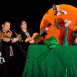 Vier Männer treten auf; zwei spielen Instrumente (Geige, Posaune), einer spielt Gitarre, und einer interagiert mit zwei Puppen auf einer grünen Bühne mit orangefarbenem Hintergrund.