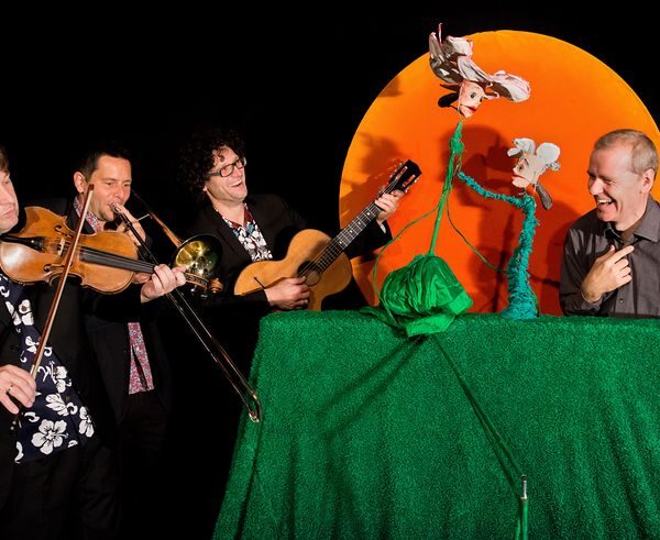 Vier Männer treten auf; zwei spielen Instrumente (Geige, Posaune), einer spielt Gitarre, und einer interagiert mit zwei Puppen auf einer grünen Bühne mit orangefarbenem Hintergrund.