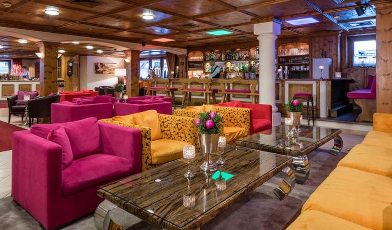 Farbenfroher Lounge-Bereich mit rosafarbenen und gelben Sesseln, gläsernen Couchtischen und einer Bar im Hintergrund, deren Decke und Wände holzvertäfelt sind.