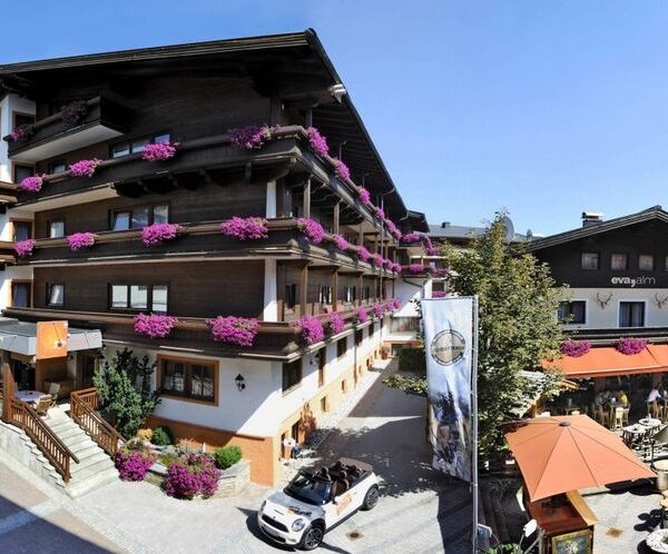 Ein mehrstöckiges Alpenhotel mit Holzbalkonen, die mit violetten Blumen geschmückt sind, mit Sitzgelegenheiten im Freien und einem Cabrio, das an einem sonnigen Tag in der Nähe parkt.