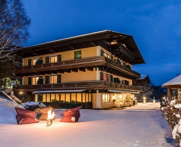 Ein dreistöckiges Gebäude im alpinen Stil mit Balkonen ist in der Abenddämmerung beleuchtet, umgeben von Schnee; ein kleines Feuer mit Sitzgelegenheiten und ein Pavillon stehen im verschneiten Hof.