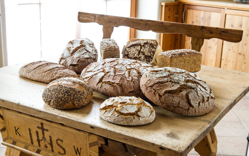 Auf einem rustikalen Holztisch in einem sonnendurchfluteten Raum mit Holzmöbeln liegen mehrere Brote mit verschiedenen Sorten aus.