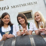 Drei Frauen in traditioneller Kleidung halten Gläser mit Weißwein in der Hand, lächeln und posieren vor einem Gebäude mit der Aufschrift „Mostmuseum“.